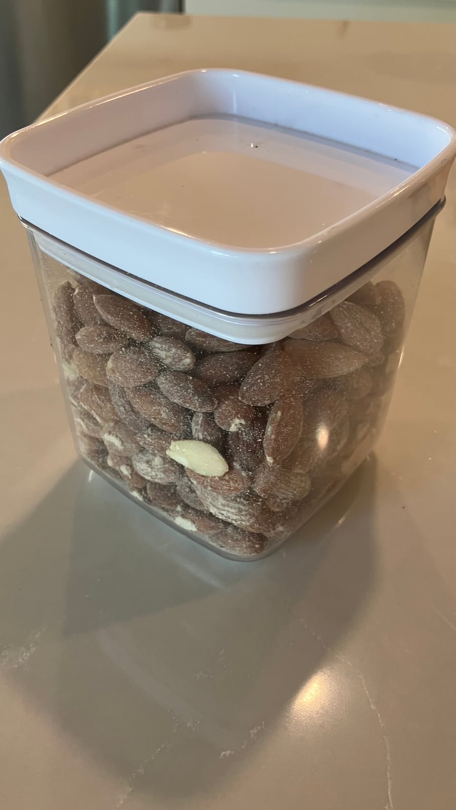 A clear square container of almonds with its white lid on, on a kitchen counter